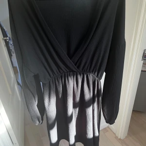 Svart omlottklänning - Snygg svart omlottklänning med v-ringning och långa ärmar. Klänningen har en elastisk midja som ger en smickrande passform. Perfekt för både vardag och festliga tillfällen.