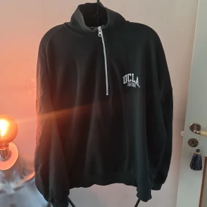 Svart tröja med dragkedja från H&M - Säljer en svart tröja från H&M med dragkedja och trycket 'UCLA' på bröstet. Tröjan har en hög krage och långa ärmar, perfekt för en avslappnad stil. Passar bra till både jeans och joggers.