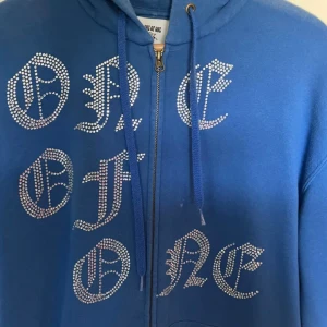 Blå hoodie med strassdetaljer från One of One - Priset kan diskuteras.                                                                Säljer en blå hoodie från One of One med texten 'ONE OF ONE' i strass på framsidan. Hoodien har dragkedja och justerbara snören vid huvan. Perfekt för en unik och stilren look.