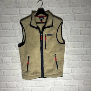 Beige fleeceväst från Patagonia - Säljer en beige fleeceväst från Patagonia med dragkedja och kontrasterande mörkblå detaljer. Västen har praktiska fickor med dragkedjor och en bekväm passform. Perfekt för lager-på-lager under kyliga dagar.