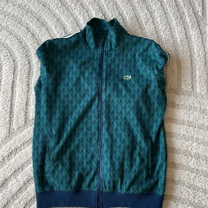 Grön tracksuit från Lacoste - Retroinspirerad grön tracksuit från Lacoste med dragkedja framtill, ståkrage och vita ränder längs ärmarna. Tracksuiten har ett diskret rutmönster och den klassiska krokodilloggan. Marinblå muddar vid ärmslut och nederkant.
