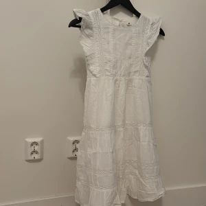 Vit klänning från H&M - Söt vit klänning från H&M med spetsdetaljer och volanger vid ärmarna. Perfekt för sommarens alla tillfällen. Klänningen har en luftig passform och knäppning i nacken.