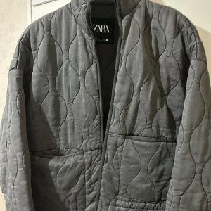 Grå quiltad jacka från Zara - Säljer en stilren grå quiltad jacka från Zara. Jackan har en dragkedja framtill och två praktiska fickor. Den är perfekt för kyligare dagar och har en modern look med sitt quiltade mönster.