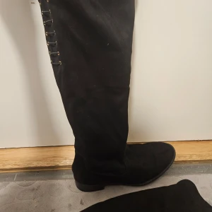 Svarta höga stövlar med snörning - Snygga svarta stövlar i mocka med snörning längs sidan. De är knähöga och har en rund tå. Perfekta för en stilren look.