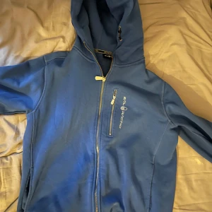 Blå hoodie från Sail Racing - Säljer en blå hoodie från Sail Racing med dragkedja och huva. Den har en bröstficka med dragkedja och texten 'Sail Racing EST. 1977' på ryggen. Perfekt för en avslappnad stil. Limited edition