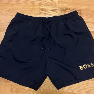 Mörkblå badshorts från Boss - Snygga mörkblå badshorts från Boss med elastisk midja och snörning. Dom är knappast använda, dom har använts några gånger då när jag badat. Guldet är av på ena snöret som man kan se på bilden, allt annat är perfekt.
