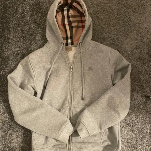 Grå Burberry Hoodie - Tja säljer denna riktigt feta burberry hoodien med monogram i luvan. Storleken är M men passar även dig som bär S. Säljer då den inte kommer till användning längre. Tröjan är i riktigt fint skick, tveka inte på att höra av dig om du har några fler frågor!