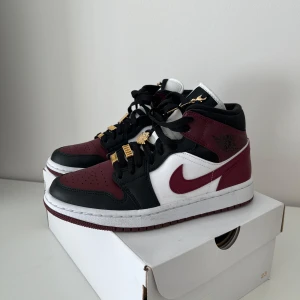 Nike Air Jordan 1 Mid SE i svart och mörkröd - Snygga Nike Air Jordan 1 Mid SE sneakers i svart, vinröd och vit. Skorna har en klassisk design med snörning och gulddetaljer på skosnörena. Endast testade utomhus. Ingen creasing. Box följer med. 
