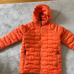 Orange dunjacka från Peak Performance - Säljer en snygg orange dunjacka från Peak Performance. Jackan har en dragkedja framtill och praktiska fickor med dragkedjor. Perfekt för kyliga dagar med sin varma och bekväma design. Huvan ger extra skydd mot väder och vind.