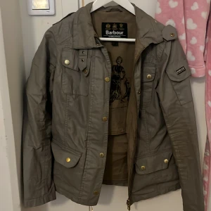 Olivgrön/grå jacka från Barbour - Säljer en olivgrön jacka från Barbour med dragkedja och knappar. Jackan har flera fickor och en klassisk design med krage och axelklaffar. Perfekt för en stilren look. Figursydd👌🏼