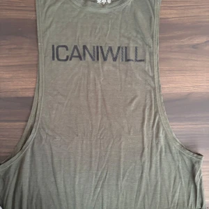 Olivgrön linnetopp från ICANIWILL - Säljer en olivgrön linnetopp från ICANIWILL. Perfekt för träning med sitt ärmlösa och luftiga design. Toppen har ett tryck med texten 'ICANIWILL' på framsidan.