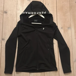 Svart huvtröja från Peak Performance - Säljer en stilren svart huvtröja från Peak Performance med dragkedja framtill. Tröjan har en justerbar huva med logotyptryck. Som ny använd en gång. 