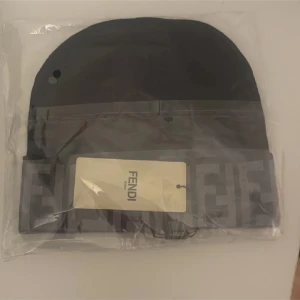 Svart mössa från Fendi - Snygg svart mössa från Fendi med det ikoniska Fendi-mönstret i grått runt kanten. Perfekt för att hålla sig varm med stil. Mössan är förpackad i plast och har en etikett med Fendi-loggan.