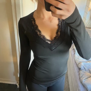 Svart långärmad topp med spetsdetaljer - Snygg svart långärmad topp med v-ringad spetsdetalj runt halsen. Perfekt för en stilren look. Passar bra till både jeans och kjol.