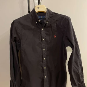 Svart skjorta från Ralph Lauren - Snygg svart skjorta från Ralph Lauren i slim fit. Skjortan har långa ärmar och knappar framtill. Den är dekorerad med Ralph Laurens ikoniska logga i rött på bröstet.