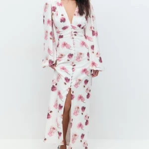 Vit blommig långklänning - Elegant vit långklänning med rosa blommönster och djup v-ringning. Klänningen har långa ärmar och en slits framtill som ger en stilfull touch. Perfekt för sommarens festligheter.
