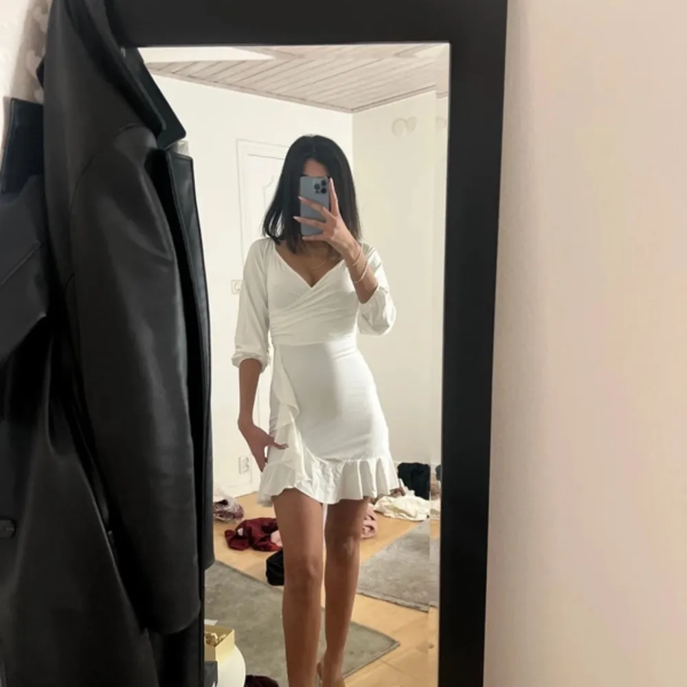 Snygg vit klänning med v-ringning och volangdetaljer vid fållen. Klänningen har trekvartsärmar och en omlott-design som ger en elegant look. Perfekt för sommarens festligheter!. Mekot.