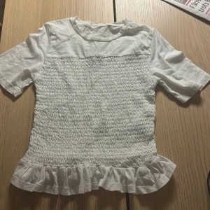 Vit topp med volangdetalj från Gina tricot💕 - Säljer en söt vit topp med smockad framsida och volangdetalj vid nederkanten. Toppen har korta ärmar. Perfekt för en somrig look!💕💕