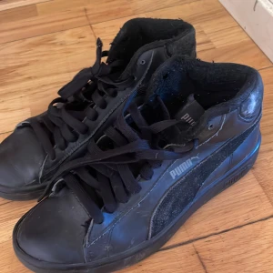 Svarta sneakers från Puma - Snygga svarta sneakers från Puma med snörning och högre skaft. Skorna har en stilren design med Puma-loggan på sidan och en robust sula för bra grepp. Perfekta för en casual look.