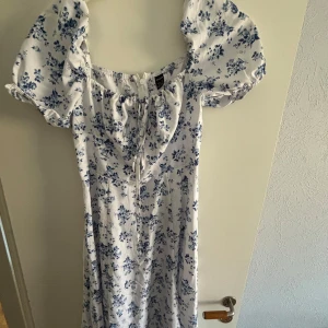 Vit blommig klänning med puffärmar - Charmig vit klänning med blått blommönster och puffiga kortärmar. Klänningen har en söt knytning framtill och en dragkedja bak för enkel påklädning. Perfekt för somriga dagar!