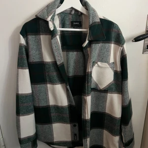 Rutig overshirt från Pull&Bear - Säljer en snygg rutig overshirt från Pull&Bear i grönt och vitt. Jackan har en klassisk krage och knäppning framtill. Perfekt för lager-på-lager-stilen. Den har en bröstficka och är tillverkad i polyester för en mjuk känsla.