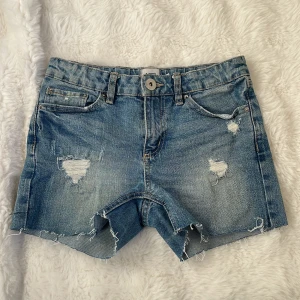 Blå jeansshorts med slitningar - Snygga blå jeansshorts med coola slitningar och fransiga kanter. Perfekta för en avslappnad stil. De har en knapp och dragkedja framtill samt klassiska fem fickor.