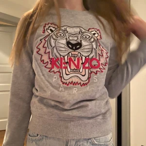 Grå sweatshirt från Kenzo med tigertryck - Säljer en grå sweatshirt från Kenzo med ett coolt tigertryck i rosa och svart. Tröjan har rund hals och långa ärmar, perfekt för en avslappnad stil. Passar bra till jeans eller kjol. 🐯