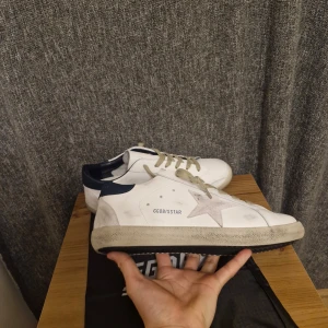 Vita sneakers från Golden Goose - Snygga vita sneakers från Golden Goose med en stjärndetalj på sidan. Skorna har grå snörning och en mörkblå häl. Perfekta för en stilren look i stl 42 priset är inte hugget i sten.