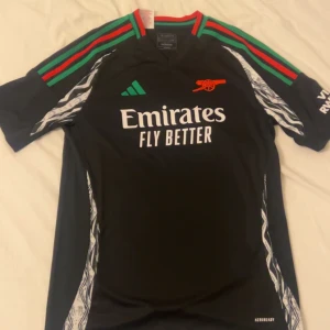 Svart fotbollströja från Adidas - Säljer en svart fotbollströja från Adidas med gröna och röda detaljer. Tröjan har korta ärmar och ett tryck med texten 'Emirates Fly Better' på framsidan. På baksidan finns namnet 'Saka' och nummer 7. Perfekt för fotbollsfans!