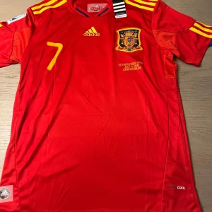 Röd fotbollströja från Adidas - Säljer en röd fotbollströja från Adidas med gula detaljer och nummer 7 på framsidan. Tröjan har korta ärmar och ett emblem på bröstet. Perfekt för fotbollsfans!