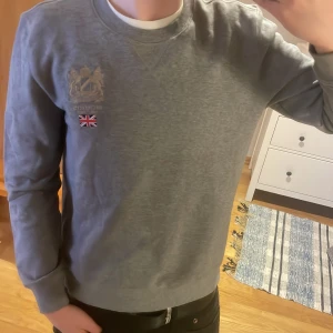 Grå sweatshirt från Morris - Säljer en stilren grå sweatshirt från Morris med broderad logga och flagga på bröstet. Tröjan har långa ärmar och rund halsringning. Perfekt för en avslappnad stil.