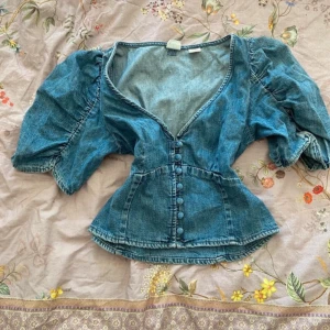 Blå jeansjacka från Levi's - Säljer en snygg blå jeansjacka från Levi's med puffiga trekvartsärmar och knappar framtill. Jackan har en v-ringad design och en figursydd passform som ger en cool look. Perfekt för att ge din outfit en extra edge!