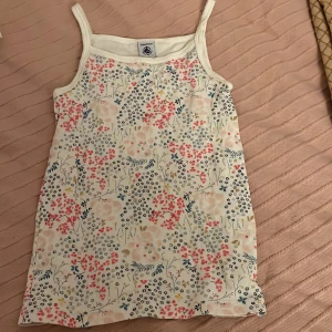 Blommig linnetopp från Petit Bateau - Säljer en söt blommig linnetopp från Petit Bateau. Toppen har ett färgglatt blommönster i rosa, blått och gult på en vit bakgrund. Perfekt för varma sommardagar!