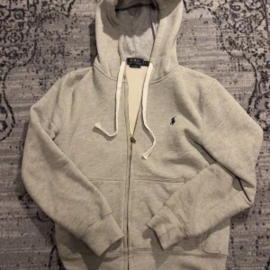 Grå hoodie från Polo Ralph Lauren - Säljer en stilren grå hoodie från Polo Ralph Lauren med dragkedja och det klassiska broderade logotypen i blått på bröstet. Tröjan har långa ärmar och en justerbar huva med vita snören. Perfekt för en avslappnad look. Skick 8/10 knappast använd! Frågor? DM!