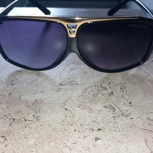 Solglasögon  - Snygga solglasögon med en elegant svart och guld design. Glasögonen har en klassisk aviatorform med mörka linser och en subtil logotyp på sidan. Perfekta för en stilren look.