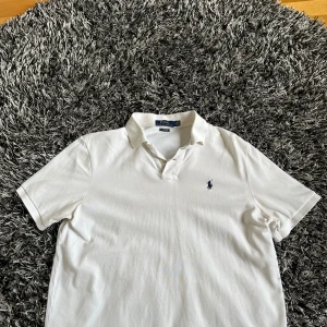 Vit pikétröja från Ralph Lauren - Säljer en klassisk vit pikétröja från Ralph Lauren med den ikoniska loggan broderad på bröstet. Tröjan har korta ärmar och en stilren krage. Perfekt för en avslappnad men ändå stilfull look. Skick: 10/10, man kan inte se att den är använd