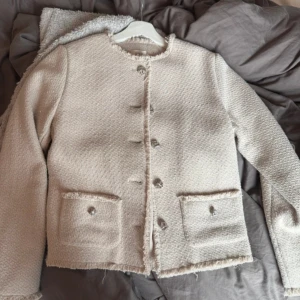 Beige kavaj från Second Female - Säljer en elegant beige kavaj från Second Female i storlek XS. Kavajen har en texturerad yta med fransiga kanter och knäpps med stiliga knappar framtill. Perfekt för en chic look!