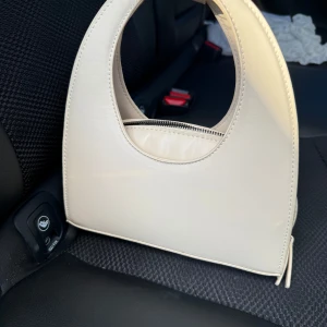 Beige handväska med dragkedja - Snygg beige handväska i syntetmaterial med en unik rundad form. Väskan har en praktisk dragkedja och ett stilrent handtag. Perfekt för att bära med sig det viktigaste med stil.