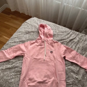 Rosa hoodie från GANT - Säljer en snygg rosa hoodie från GANT med justerbar dragsko i huvan och en praktisk magficka. Perfekt för en avslappnad stil. GANT-loggan är broderad på bröstet.