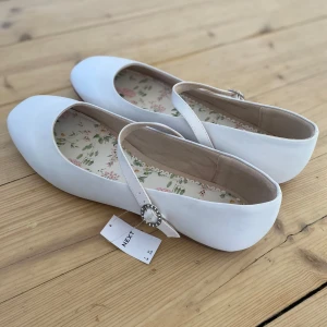 Vita ballerinaskor  - Säljer ett par vita ballerinaskor från Next med en dekorativ rem och glittrande spänne. Perfekta för studenten. Aldrig använda. 