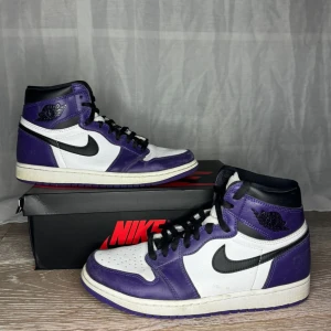 Jordan 1 Court Purple - Snygga Nike Air Jordans i en cool lila och vit färgkombination. Skorna har en klassisk hög design med svart snörning och Nike-logga på sidan. Perfekta för att ge din outfit en sportig touch.