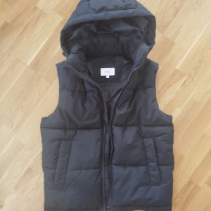 Svart quiltad dunväst med huva - Snygg svart quiltad dunväst med dragkedja och avtagbar huva. Köpt i Frankrike och bara använd 3 gånger. Västen är av storlek S som i sverige borde motsvara ca storlek 46. Västen har praktiska fickor och en stilren design.