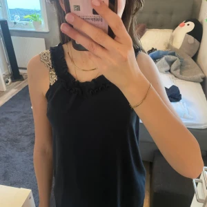 Svart ärmlös topp med volangdetaljer - Snygg svart ärmlös topp med volangdetaljer vid axlarna. Perfekt för en stilren look. Toppen har en enkel design som gör den lätt att matcha med olika outfits.