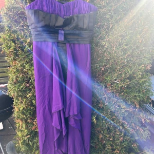 Festklänning  - Säljer en elegant lila axelband klänning med svart banddetalj i midjan. Klänningen har en vacker volangdesign och är perfekt för speciella tillfällen. Den har en lång, flytande kjol som ger en dramatisk look. 