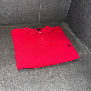 Röd pikétröja från Ralph Lauren - Säljer en klassisk röd pikétröja från Ralph Lauren. Tröjan är tillverkad i 100% bomull och har en broderad logotyp på bröstet. Den har korta ärmar och en traditionell krage med knappar. Perfekt för en stilren look. Modellen i bilden är 174 cm