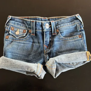 Jeansshorts från True Religion - Snygga jeansshorts från True Religion med klassisk femficksdesign och uppvikta ben. De har dekorativa sömmar och en ikonisk logga på bakfickan. Perfekta för en avslappnad stil.