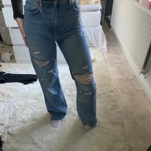 Blå jeans med slitningar - Snygga blå jeans med hög midja och slitningar på framsidan. De har en klassisk femficksdesign och en straight passform som ger en avslappnad look. Perfekta för en trendig vardagsstil.