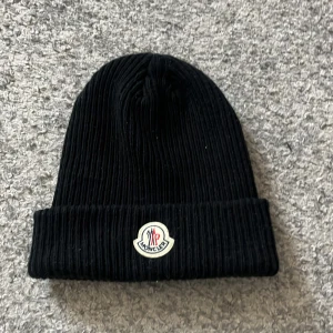 Svart ribbad mössa från Moncler - Snygg svart ribbad mössa från Moncler med deras ikoniska logotyp framtill. Perfekt för att hålla värmen under kyliga dagar. Stilren och enkel design som passar till det mesta.