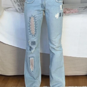 Ljusblå jeans med patchwork och slitningar - Unika ljusblå jeans med coola patchwork-detaljer och slitningar. Byxorna har en straight passform och är dekorerade med kontrasterande sömmar och lappar. Perfekta för en edgy look! Midjemått: 39cm och innerbenslängd: 79 cm