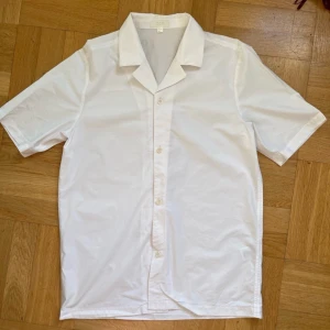 Vit kortärmad skjorta från COS - Säljer en stilren vit kortärmad skjorta från COS. Skjortan har en klassisk krage och knappar framtill. Perfekt för en minimalistisk look.